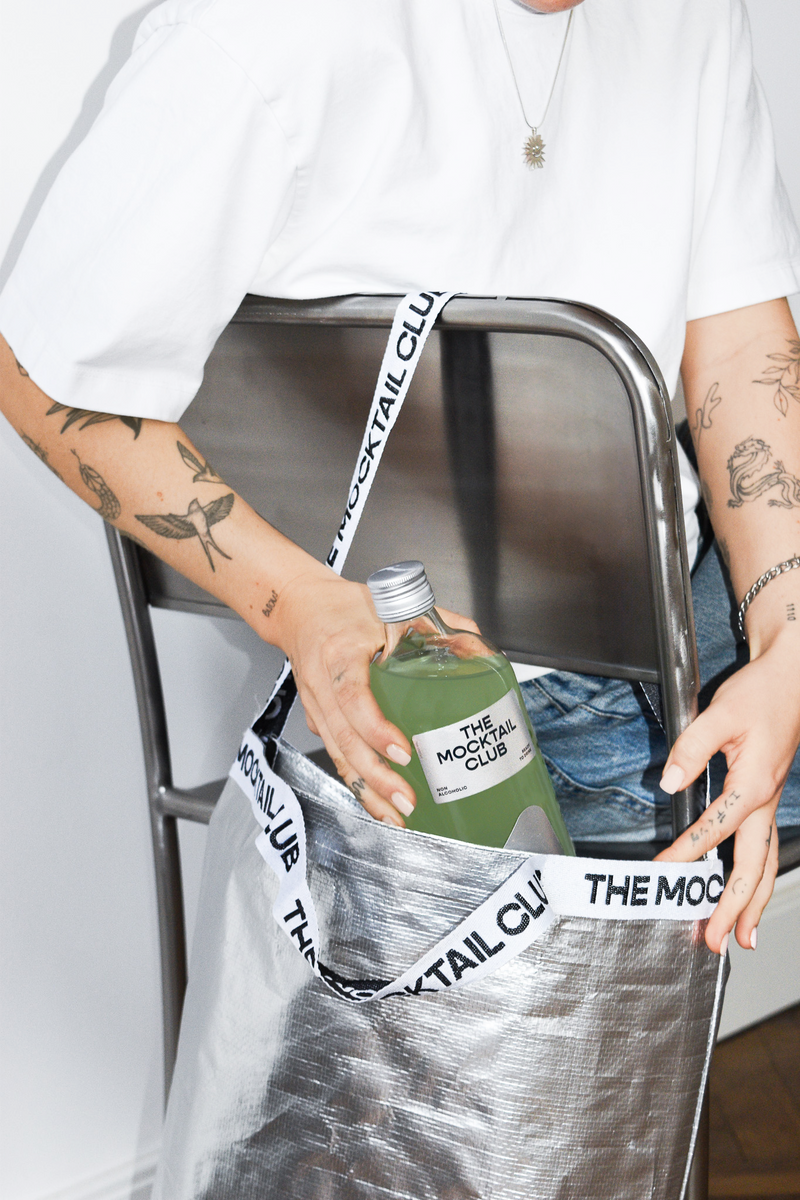 Woman putting a bottle of Basil Breeze in a  silver The Mocktail Club shopping bag 